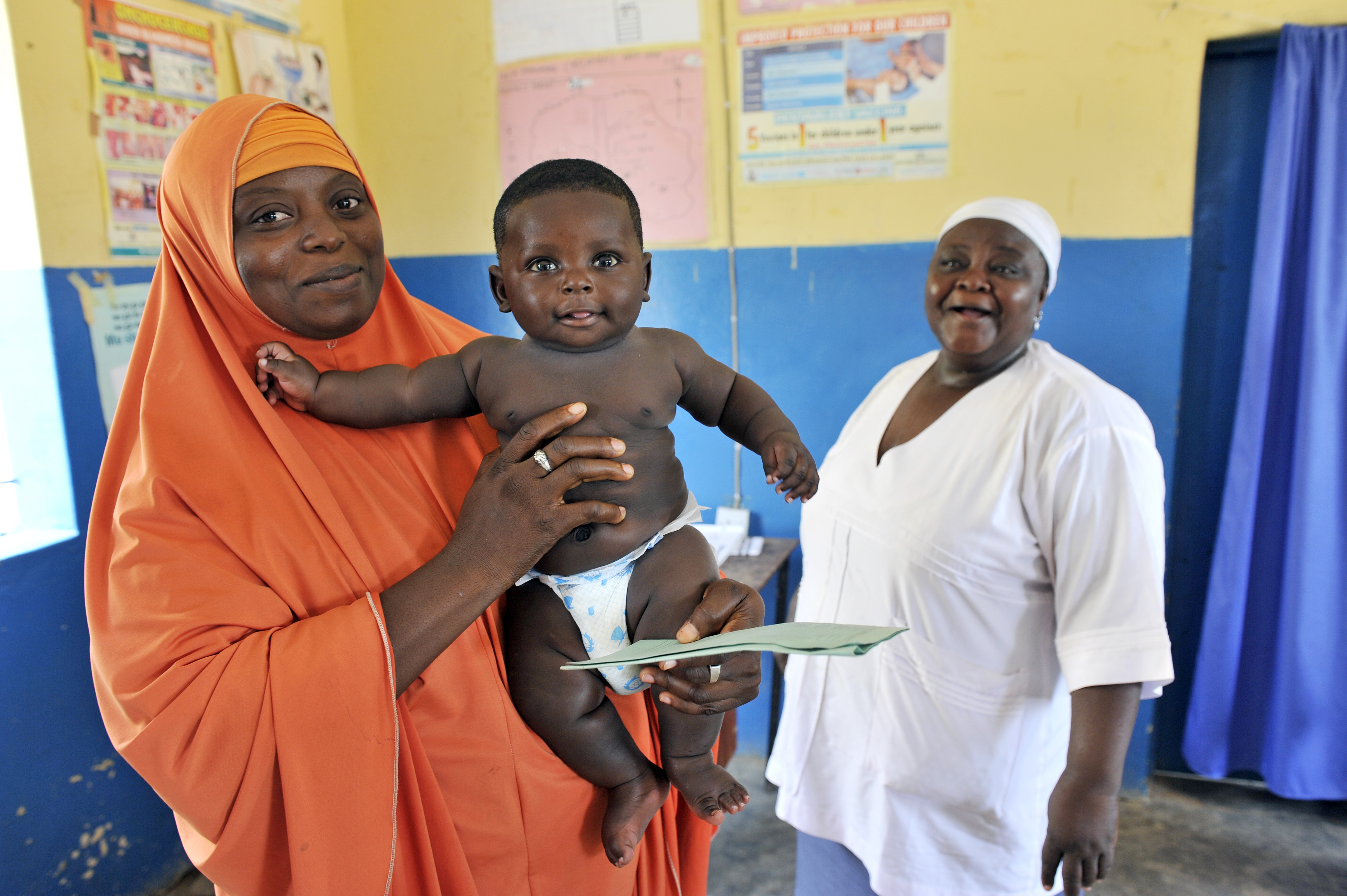 A mother in Nigeria holds her infant in a clinical setting. A nurse looks on, smiling, in the background.